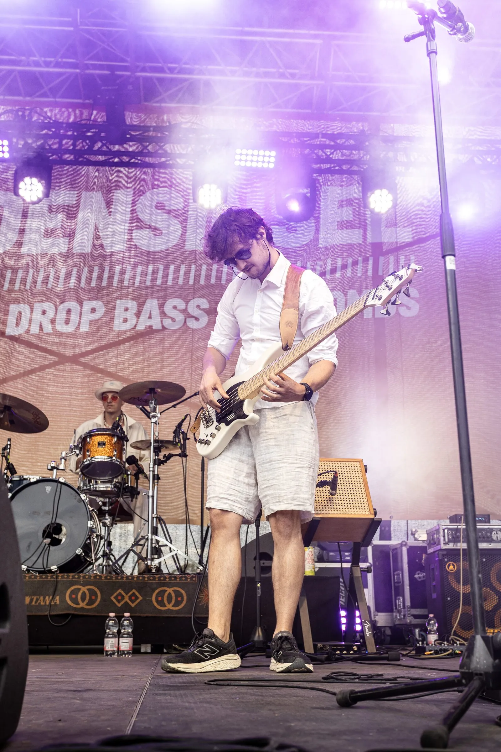 Bass in Weiß unter violettem Licht, dahinter das Schlagzeug