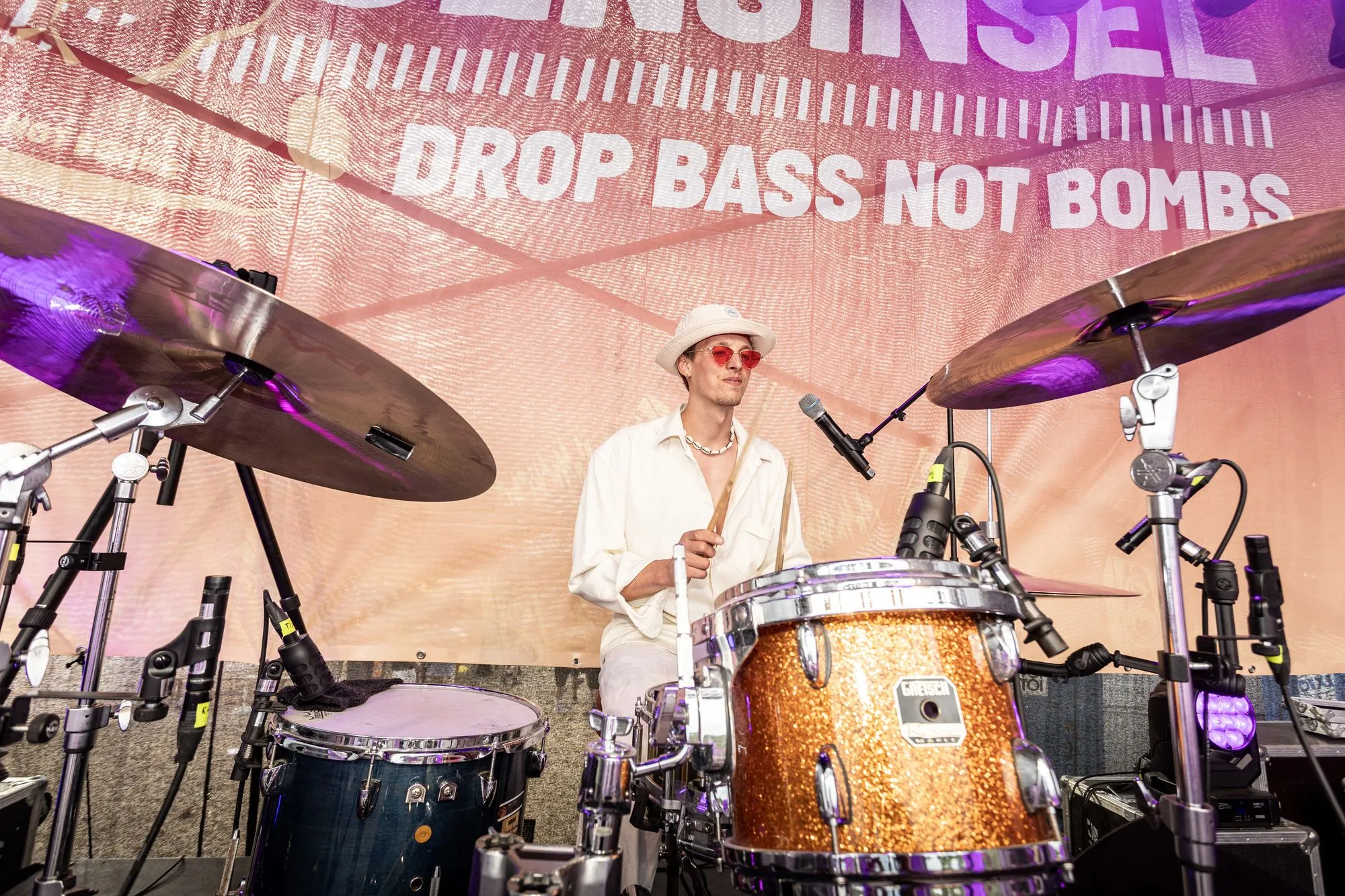 Am Schlagzeug mit weißem Hut, dahinter „Drop Bass Not Bombs“